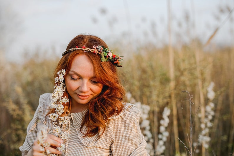 May include: A woman with long, red hair wears a floral crown and a light brown, checkered dress. She is holding a sprig of white flowers in her hand and looking down at them.