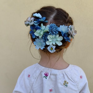 K&ouml;nnte beinhalten: Ein Kind mit dunklem Haar, das ein wei&szlig;es Cordhemd mit floraler Stickerei tr&auml;gt. Ein Blumenkranz aus blauen und hellblauen Blumen schm&uuml;ckt das Haar und erzeugt einen zarten Look. Der Hintergrund ist einfarbig hellgelb.
