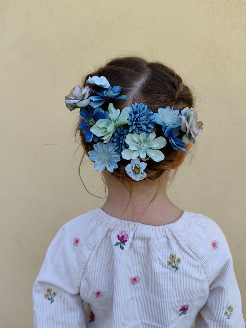 K&ouml;nnte beinhalten: Ein Kind mit dunklem Haar, das ein wei&szlig;es Cordhemd mit floraler Stickerei tr&auml;gt. Ein Blumenkranz aus blauen und hellblauen Blumen schm&uuml;ckt das Haar und erzeugt einen zarten Look. Der Hintergrund ist einfarbig hellgelb.