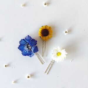 May include: Three gold hair clips with different flower designs. A blue flower, a yellow sunflower, and a white daisy.