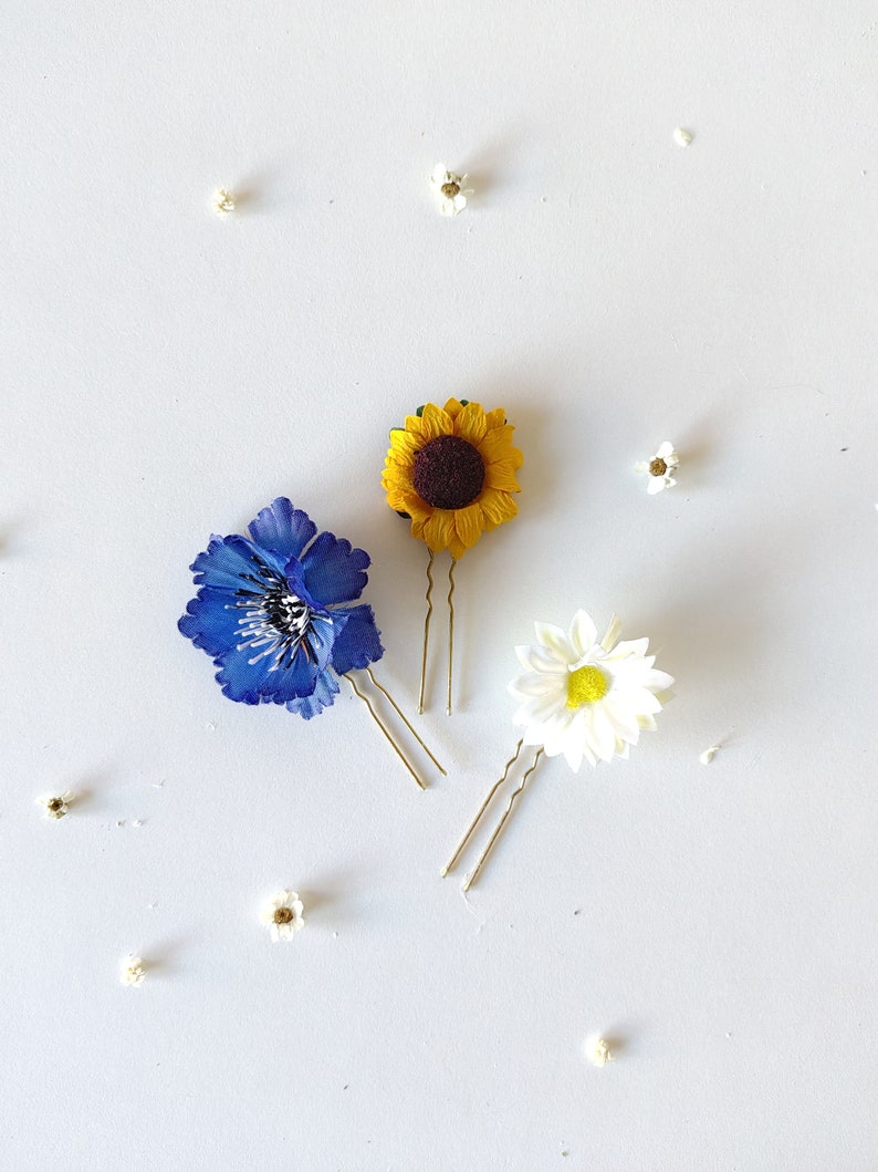 May include: Three gold hair clips with different flower designs. A blue flower, a yellow sunflower, and a white daisy.