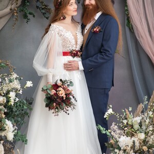 May include: A bride and groom stand in front of a floral backdrop. The bride is wearing a white wedding dress with a red sash and a floral crown. The groom is wearing a blue suit with a brown vest. They are both holding bouquets of flowers.