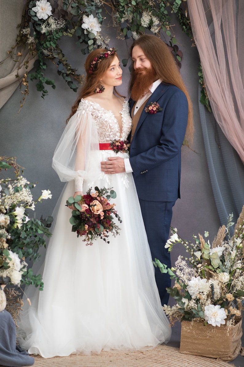 May include: A bride and groom stand in front of a floral backdrop. The bride is wearing a white wedding dress with a red sash and a floral crown. The groom is wearing a blue suit with a brown vest. They are both holding bouquets of flowers.