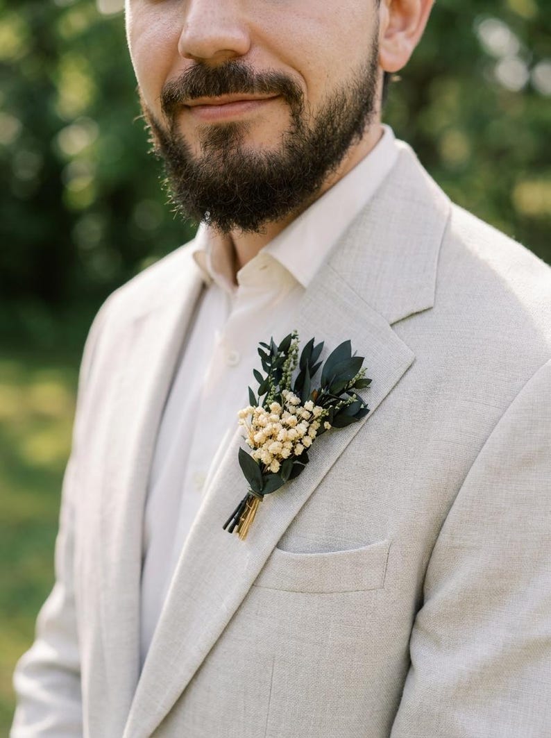May include: A light grey blazer with a white shirt and a floral buttonhole. The buttonhole features small white flowers, dark green leaves, and gold accents. The blazer has a classic lapel and a pocket.