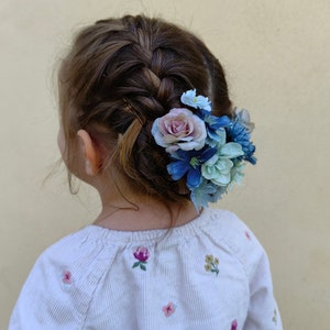 K&ouml;nnte beinhalten: Geflochtenes Haar einer jungen Person, verziert mit einem Cluster aus blauen, hellblauen und rosa Kunstblumen. Die Person tr&auml;gt ein wei&szlig;es Langarmshirt mit gestickten floralen Details.