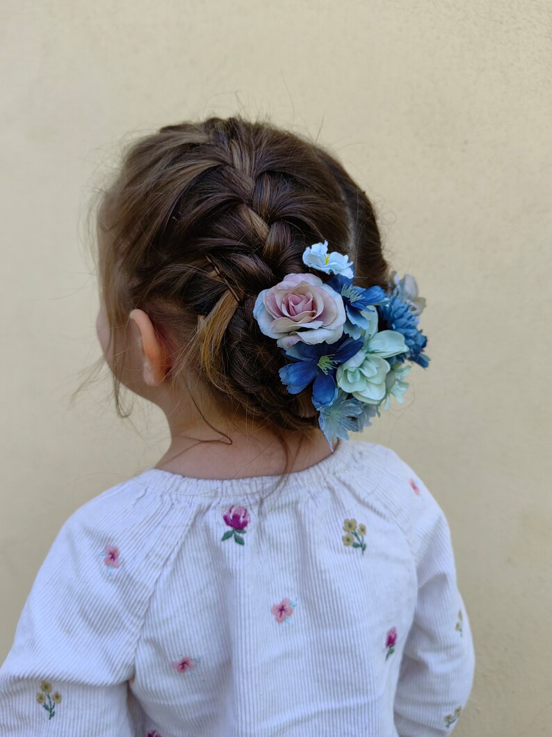 K&ouml;nnte beinhalten: Geflochtenes Haar einer jungen Person, verziert mit einem Cluster aus blauen, hellblauen und rosa Kunstblumen. Die Person tr&auml;gt ein wei&szlig;es Langarmshirt mit gestickten floralen Details.