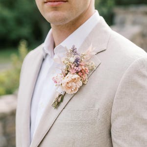 May include: A beige blazer with a white shirt and a floral boutonniere. The boutonniere features a large peach-colored flower, smaller pink and purple flowers, and greenery, tied with lace.