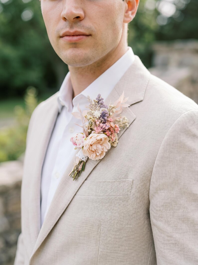 May include: A beige blazer with a white shirt and a floral boutonniere. The boutonniere features a large peach-colored flower, smaller pink and purple flowers, and greenery, tied with lace.
