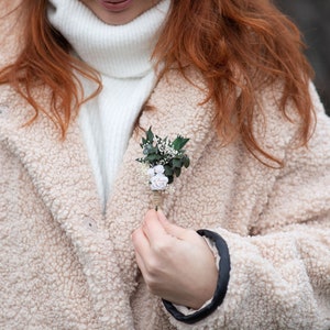 Puede incluir: Un peque&ntilde;o prendedor floral con detalles blancos y verde claro est&aacute; prendido a un abrigo de peluche beige. El prendedor presenta peque&ntilde;as rosas blancas y vegetaci&oacute;n, sujetas con hilo. El abrigo est&aacute; abotonado sobre un cuello alto blanco.