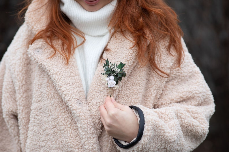 Puede incluir: Un peque&ntilde;o prendedor floral con detalles blancos y verde claro est&aacute; prendido a un abrigo de peluche beige. El prendedor presenta peque&ntilde;as rosas blancas y vegetaci&oacute;n, sujetas con hilo. El abrigo est&aacute; abotonado sobre un cuello alto blanco.
