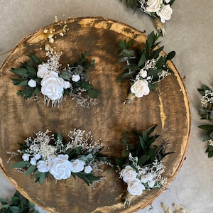Puede incluir: Una variedad de peinetas y boutonnieres florales. Las peinetas presentan rosas blancas, vegetaci&oacute;n y paniculata. Los boutonnieres tienen rosas blancas, vegetaci&oacute;n y est&aacute;n atados con hilo. Los art&iacute;culos est&aacute;n dispuestos sobre una superficie de madera.