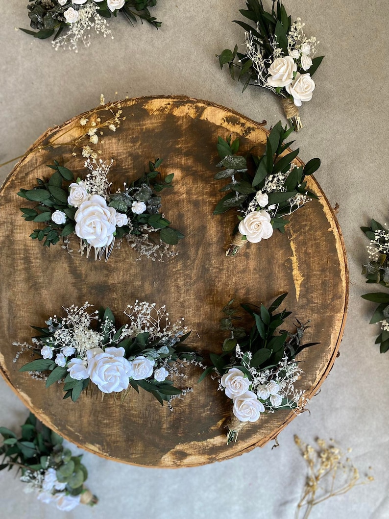 Puede incluir: Una variedad de peinetas y boutonnieres florales. Las peinetas presentan rosas blancas, vegetaci&oacute;n y paniculata. Los boutonnieres tienen rosas blancas, vegetaci&oacute;n y est&aacute;n atados con hilo. Los art&iacute;culos est&aacute;n dispuestos sobre una superficie de madera.