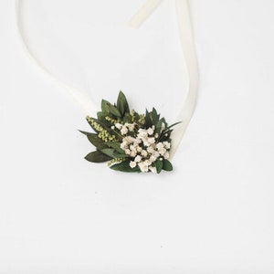 May include: A floral hair accessory with small white flowers, green leaves, and sprigs. The arrangement is attached to a white ribbon. The design is delicate and natural. Ideal for weddings or special occasions.