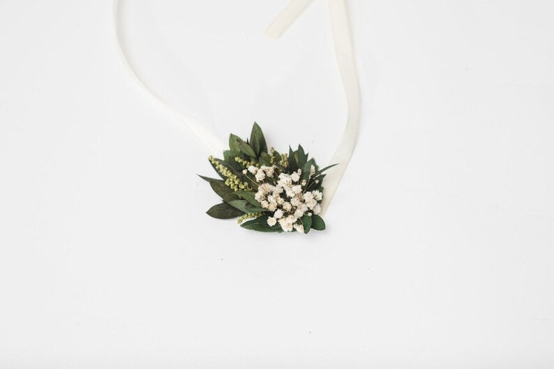 May include: A floral hair accessory with small white flowers, green leaves, and sprigs. The arrangement is attached to a white ribbon. The design is delicate and natural. Ideal for weddings or special occasions.