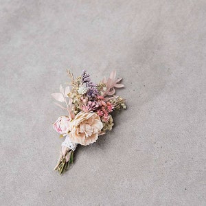 May include: A delicate floral boutonniere featuring a mix of dried flowers in soft pink, peach, and lavender hues. The arrangement includes a large, light peach peony and is wrapped with lace and twine.