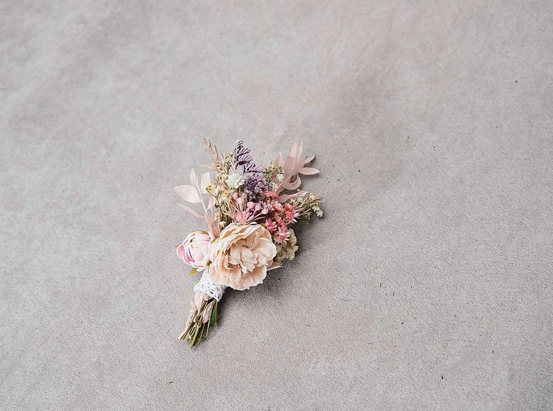 May include: A delicate floral boutonniere featuring a mix of dried flowers in soft pink, peach, and lavender hues. The arrangement includes a large, light peach peony and is wrapped with lace and twine.