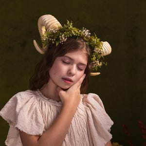 May include: A young person with long brown hair wearing a white dress and a floral crown with horns. The background is a dark green.