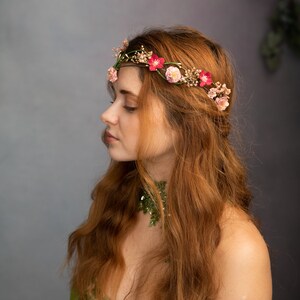 May include: A woman with long, red hair wearing a floral crown with pink and red flowers. She is wearing a green and white dress with a green leafy choker.