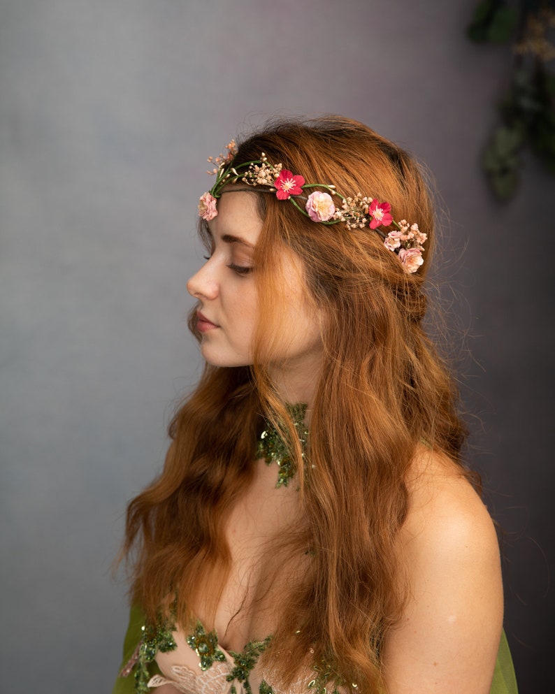 May include: A woman with long, red hair wearing a floral crown with pink and red flowers. She is wearing a green and white dress with a green leafy choker.