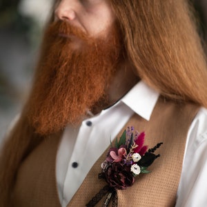 May include: A brown waistcoat with a floral buttonhole featuring burgundy, pink, and white flowers with black feathers and a black lace ribbon.