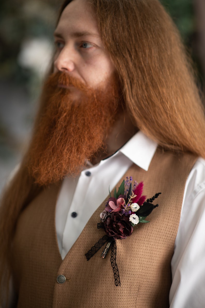 May include: A brown waistcoat with a floral buttonhole featuring burgundy, pink, and white flowers with black feathers and a black lace ribbon.