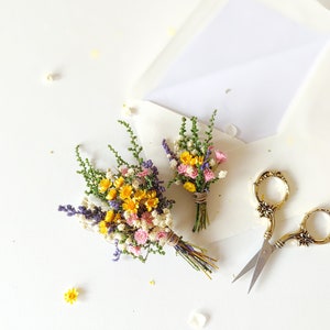 Op de afbeelding: Twee kleine boeketten gedroogde bloemen, een met gele, roze en paarse bloemen, en de andere met gele en roze bloemen, zijn vastgebonden met touw en geplaatst op een wit oppervlak. Een gouden schaar bevindt zich ook op het oppervlak.