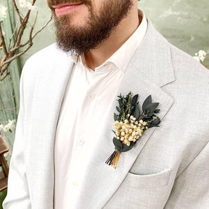 May include: A light grey blazer with a white shirt and a small floral boutonniere. The boutonniere features small white flowers, dark green leaves, and gold and black accents. The blazer has a pocket on the left side.