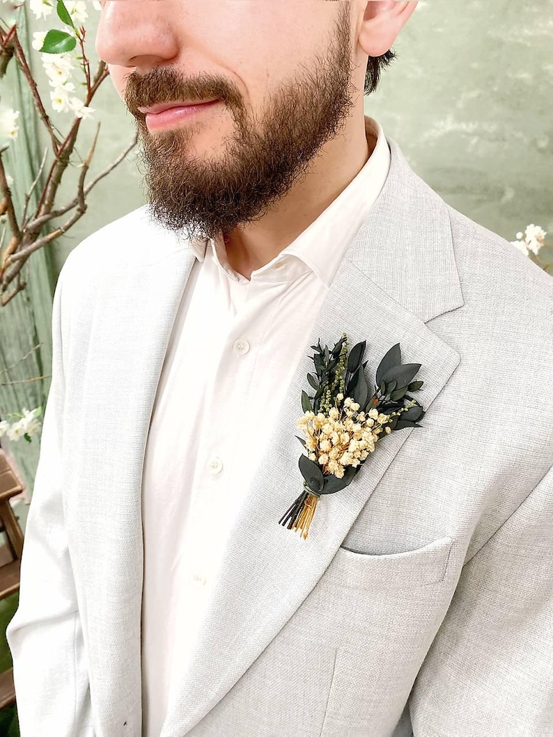 May include: A light grey blazer with a white shirt and a small floral boutonniere. The boutonniere features small white flowers, dark green leaves, and gold and black accents. The blazer has a pocket on the left side.