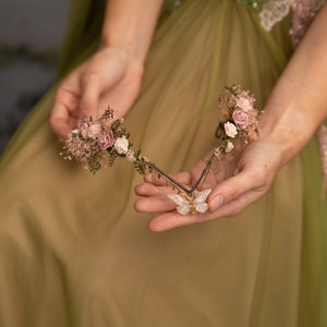 Peut inclure: Une couronne florale d&eacute;licate avec des fleurs roses et de la verdure, rehauss&eacute;e d'un charme papillon scintillant. La couronne est tenue dans les mains d'une personne portant une robe verte.