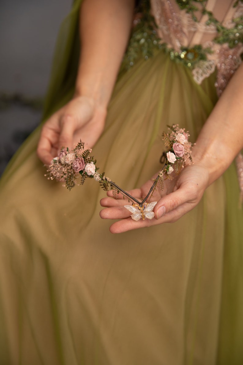 Peut inclure: Une couronne florale d&eacute;licate avec des fleurs roses et de la verdure, rehauss&eacute;e d'un charme papillon scintillant. La couronne est tenue dans les mains d'une personne portant une robe verte.