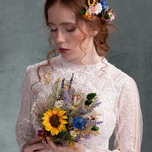 May include: A bride in a white lace dress holds a bouquet of dried flowers, including sunflowers, lavender, and wildflowers. The bouquet is held in front of her, and she is looking down at it. She is wearing a flower crown with sunflowers, daisies, and blue flowers.