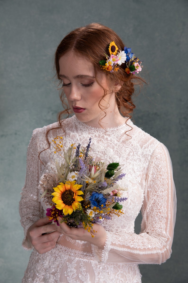 May include: A bride in a white lace dress holds a bouquet of dried flowers, including sunflowers, lavender, and wildflowers. The bouquet is held in front of her, and she is looking down at it. She is wearing a flower crown with sunflowers, daisies, and blue flowers.