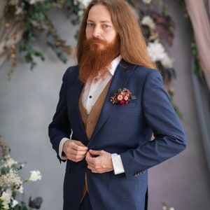 May include: A man wearing a blue suit with a brown vest and a white shirt. He has long red hair and a red beard. He is wearing a small floral boutonniere on his lapel.