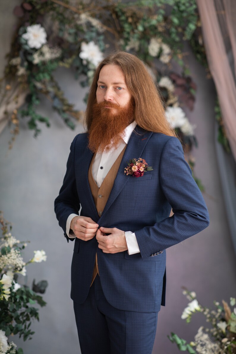 May include: A man wearing a blue suit with a brown vest and a white shirt. He has long red hair and a red beard. He is wearing a small floral boutonniere on his lapel.