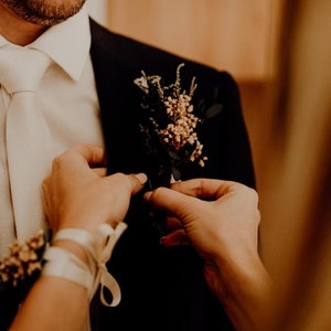May include: A person in a black suit with a white tie and a floral boutonniere. Someone is adjusting the boutonniere. A matching floral wrist corsage is also visible. The image has a warm, soft focus.