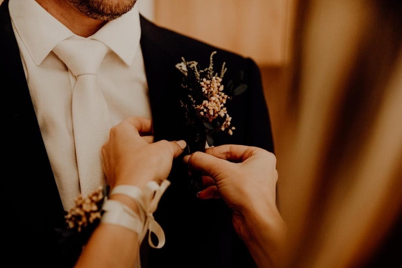 May include: A person in a black suit with a white tie and a floral boutonniere. Someone is adjusting the boutonniere. A matching floral wrist corsage is also visible. The image has a warm, soft focus.