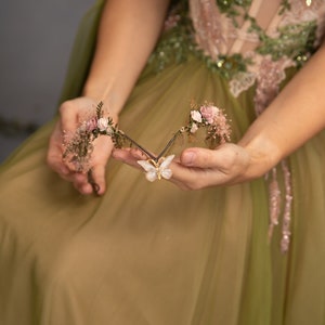 Peut inclure: Une d&eacute;licate pi&egrave;ce de cheveux florale avec un charme papillon. La pi&egrave;ce de cheveux est faite de petites fleurs roses, de feuilles vertes et d'un papillon dor&eacute;. Elle est tenue dans les mains d'une personne portant une robe verte.