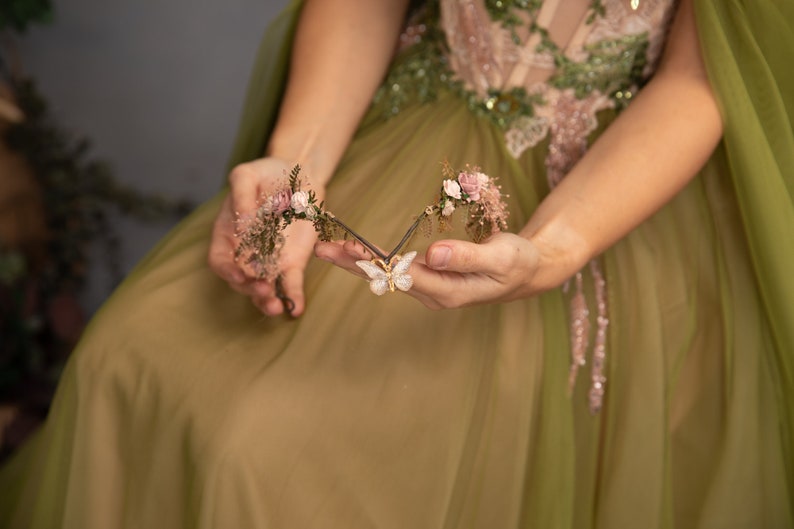 Peut inclure: Une d&eacute;licate pi&egrave;ce de cheveux florale avec un charme papillon. La pi&egrave;ce de cheveux est faite de petites fleurs roses, de feuilles vertes et d'un papillon dor&eacute;. Elle est tenue dans les mains d'une personne portant une robe verte.