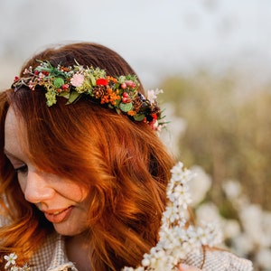 May include: A woman with long, red hair wears a floral crown with berries, pine cones, and greenery. She is wearing a light brown and white plaid shirt and is holding a sprig of white flowers.