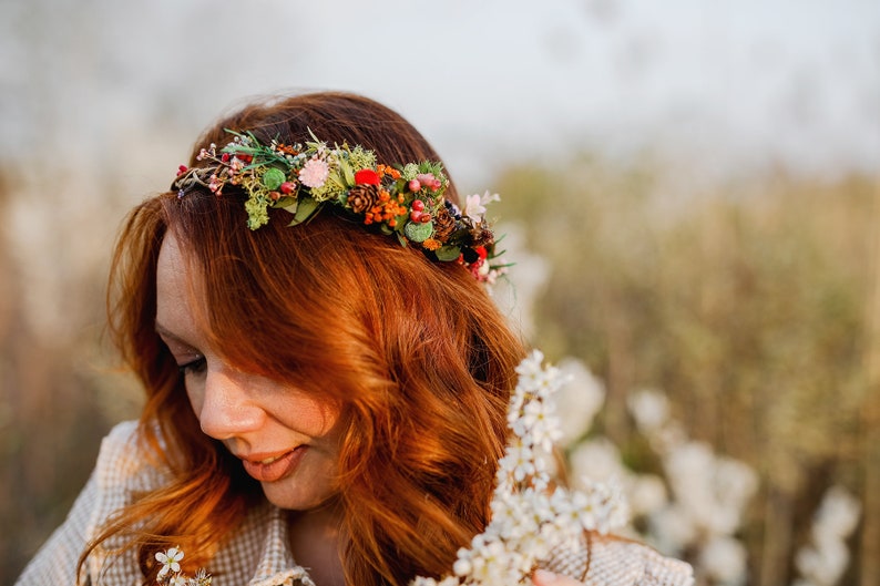 May include: A woman with long, red hair wears a floral crown with berries, pine cones, and greenery. She is wearing a light brown and white plaid shirt and is holding a sprig of white flowers.