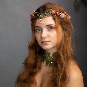 May include: A woman with long, wavy red hair wears a floral crown with pink and red flowers. She is wearing a green, sequined, off-the-shoulder dress.