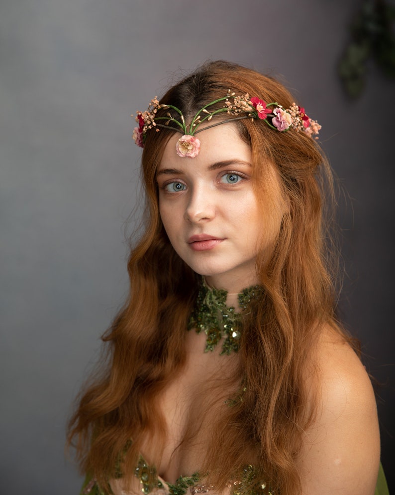 May include: A woman with long, wavy red hair wears a floral crown with pink and red flowers. She is wearing a green, sequined, off-the-shoulder dress.