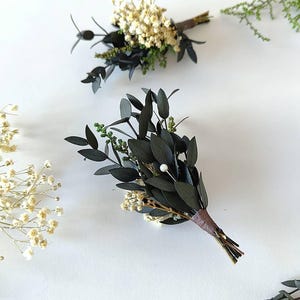May include: Two small, handcrafted boutonnieres with dark green leaves, white flowers, and a brown stem wrap. One boutonniere has a small white pearl. The boutonnieres are arranged on a white background with additional greenery.