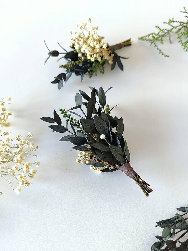 May include: Two small, handcrafted boutonnieres with dark green leaves, white flowers, and a brown stem wrap. One boutonniere has a small white pearl. The boutonnieres are arranged on a white background with additional greenery.
