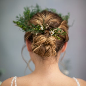 May include: A woman with blonde hair styled in a bun with a floral crown made of greenery, small white flowers, and brown pinecones.