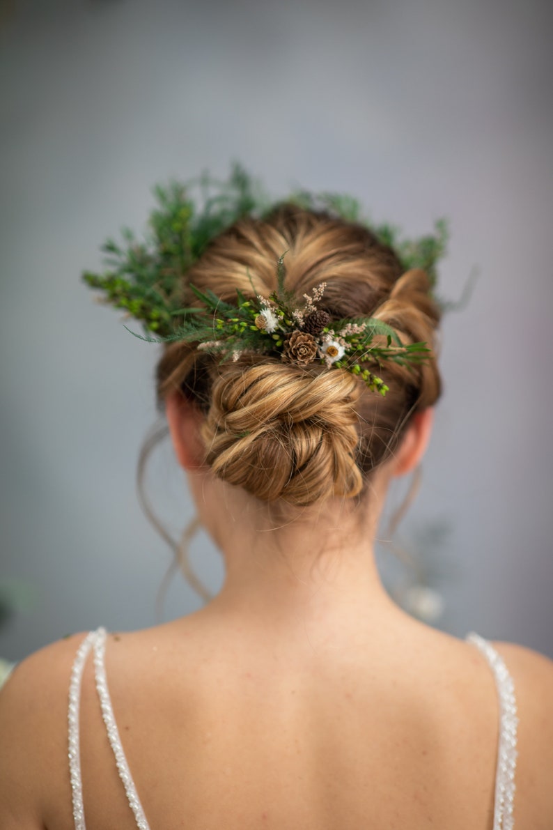 May include: A woman with blonde hair styled in a bun with a floral crown made of greenery, small white flowers, and brown pinecones.
