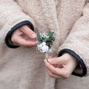 Puede incluir: Un peque&ntilde;o prendedor artesanal con rosas blancas de papel, hojas verdes y delicadas flores blancas de relleno. El arreglo floral est&aacute; envuelto con hilo y tiene un alfiler dorado. El fondo muestra un abrigo beige con ribete negro.