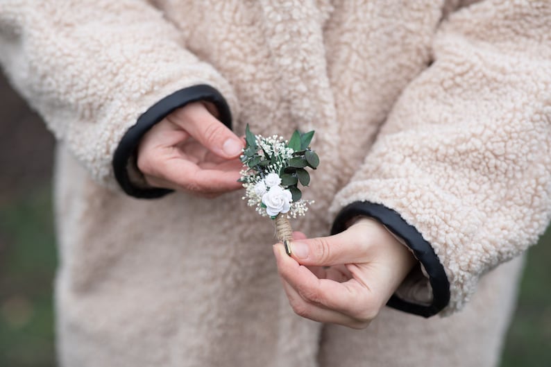 Puede incluir: Un peque&ntilde;o prendedor artesanal con rosas blancas de papel, hojas verdes y delicadas flores blancas de relleno. El arreglo floral est&aacute; envuelto con hilo y tiene un alfiler dorado. El fondo muestra un abrigo beige con ribete negro.