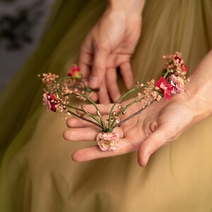 May include: A delicate floral crown with pink and red flowers, gold accents, and green leaves. The crown is held in the palm of a hand.