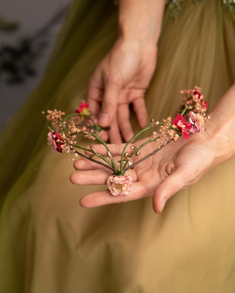 May include: A delicate floral crown with pink and red flowers, gold accents, and green leaves. The crown is held in the palm of a hand.
